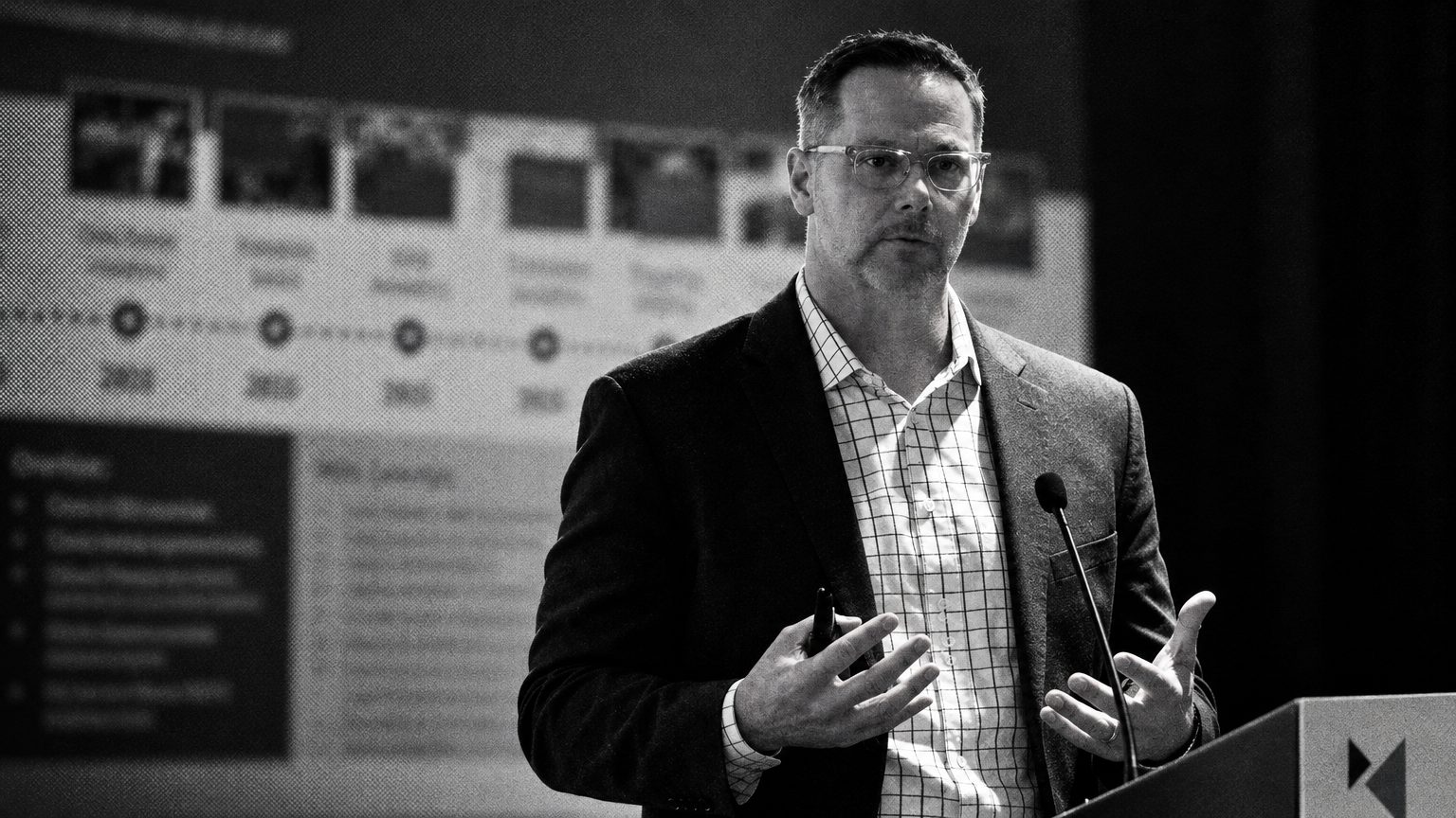 The speaker mid-gesture before a blurred timeline slide in newspaper halftone